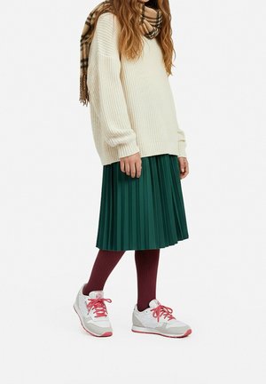Persoon die een crèmekleurige gebreide trui draagt, een groene geplooide rok, bordeauxrode maillots, witte sneakers met roze veters en een beige geruite sjaal.
