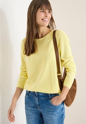 Lächelnde Frau mit gelbem Pullover und blauer Jeans, die eine braune Umhängetasche trägt und mit einer Hand in der Tasche zur Seite blickt.