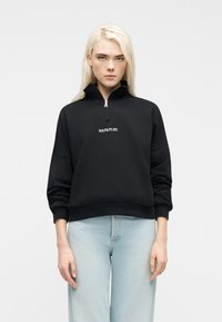 Ξανθός άνθρωπος που φοράει μαύρο φούτερ με μισή φερμουάρ και λογότυπο "NAPAPIJRI" και ανοιχτόχρωμο μπλε τζιν, στέκεται μπροστά σε απλό λευκό φόντο.