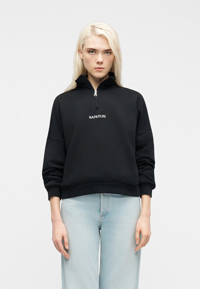 Ξανθός άνθρωπος που φοράει μαύρο φούτερ με μισή φερμουάρ και λογότυπο "NAPAPIJRI" και ανοιχτόχρωμο μπλε τζιν, στέκεται μπροστά σε απλό λευκό φόντο.