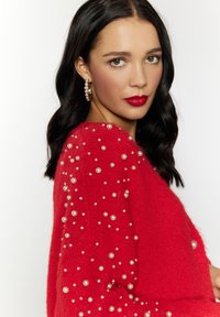 Maglione rosso con lavorazione a maglia testurizzata e perle bianche disposte a ciuffi. Il modello ha i capelli scuri e presenta labbra rosse audaci e orecchini di perle.