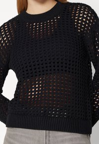 Dunkler Strickpullover mit lockerem, offenem Strickmuster, geripptem Rundhalsausschnitt und Bündchen, durch das die Haut sichtbar ist.