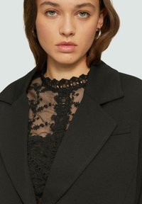 Schwarze Blazerjacke mit Reverskragen und einer einzelnen Vordertasche, überlagert mit einem schwarzen Spitzenoberteil, das mit floralen Mustern und gewellten Kanten verziert ist.