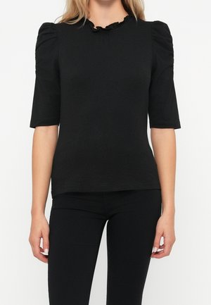 Frau trägt ein schlichtes schwarzes, figurbetontes Top mit aufgepufften kurzen Ärmeln und einem gerafften Ausschnitt, kombiniert mit schwarzen Hosen.