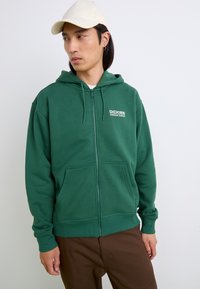 Groene ritskap die is gemaakt van zachte stof, met een voorzak, een capuchon met trekkoord en een wit logo op de borst. Gecombineerd met bruine pantalon.
