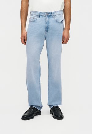 Mężczyzna ubrany w jasnoniebieskie spodnie typu straight leg oraz czarne skórzane loafersy, stojący na tle jednolitego białego tła.