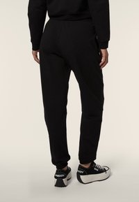 Joggers negros de elastano con un diseño ajustado. El material parece suave con una textura lisa. Llevados con zapatillas negras y blancas que presentan un logo.