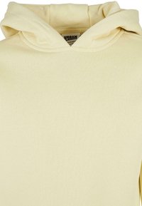 Sudadera amarilla clara con capucha ajustable, textura de tela suave y un diseño sencillo. Se ve una etiqueta con "URBAN CLASSICS" en el interior.