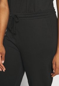 Schwarzer Stoffoverall mit gerafftem Bund und Kordelzug, seitlichen Taschen und glatter Textur, der sich auf die Taille und die Hand konzentriert.