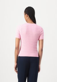 Pull rose en tricot à manches courtes avec un motif texturé et un ourlet côtelé. Porté avec un pantalon bleu marine, vu de dos.