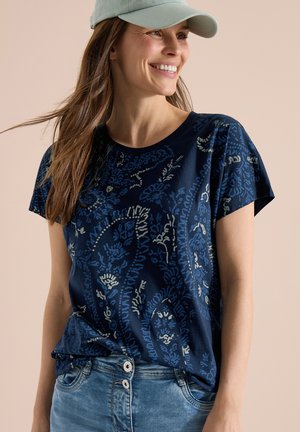 Uśmiechnięta kobieta w jasnozielonej czapce, niebieskiej koszuli w krótkim rękawie w wzory i jasno-niebieskich dżinsach z wysokim stanem.