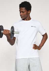 Hvid bomulds t-shirt med et sort broderet "Champion" logo på brystet, båret af en person der løfter en sort håndvægt, iført grå shorts.