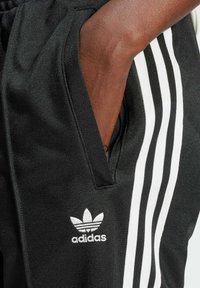 Czarne spodnie sportowe z białymi trzema paskami, teksturowana tkanina, elastyczny pas i haftowane białe logo z boku.