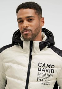 Man with short curly hair wearing a white and black puffer jacket with "CAMP DAVID CHALLENGE" text on the front, looking at the camera.