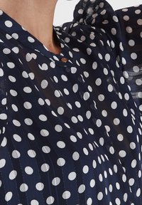 Chemisier transparent bleu marine à pois blancs, avec un col rond et une fermeture à boutons, en tissu texturé et avec des manches bouffantes.