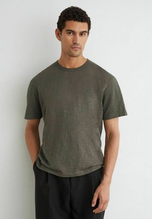 Man met donker krullend haar, gekleed in een olijfgroene korte mouwen T-shirt en een zwarte broek, staand tegen een effen lichte muur.