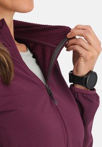 Veste zippée violette avec doublure texturée, présentant un col montant et des poignets élastiques. Une montre intelligente est visible au poignet.