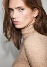 Maglione beige a coste con collo alto e dettagli di leggera lucentezza. I lunghi capelli castani e orecchini a cerchio completano il look. Sfondo neutro.