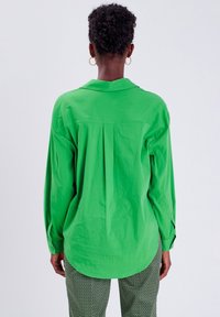 Zielona koszula z długim rękawem i kołnierzykiem, luźny krój, z detalem plisowania na plecach. Gładki materiał, zaokrąglony dół, w zestawie ze wzorzystymi spodniami.