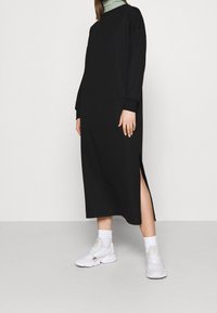 Musta midi-mekko, jossa on pitkät hihat, sivureiät ja rento istuvuus, yhdistettynä valkoisiin tennareihin ja joustaviin sukkiin. Sujuva kangas.