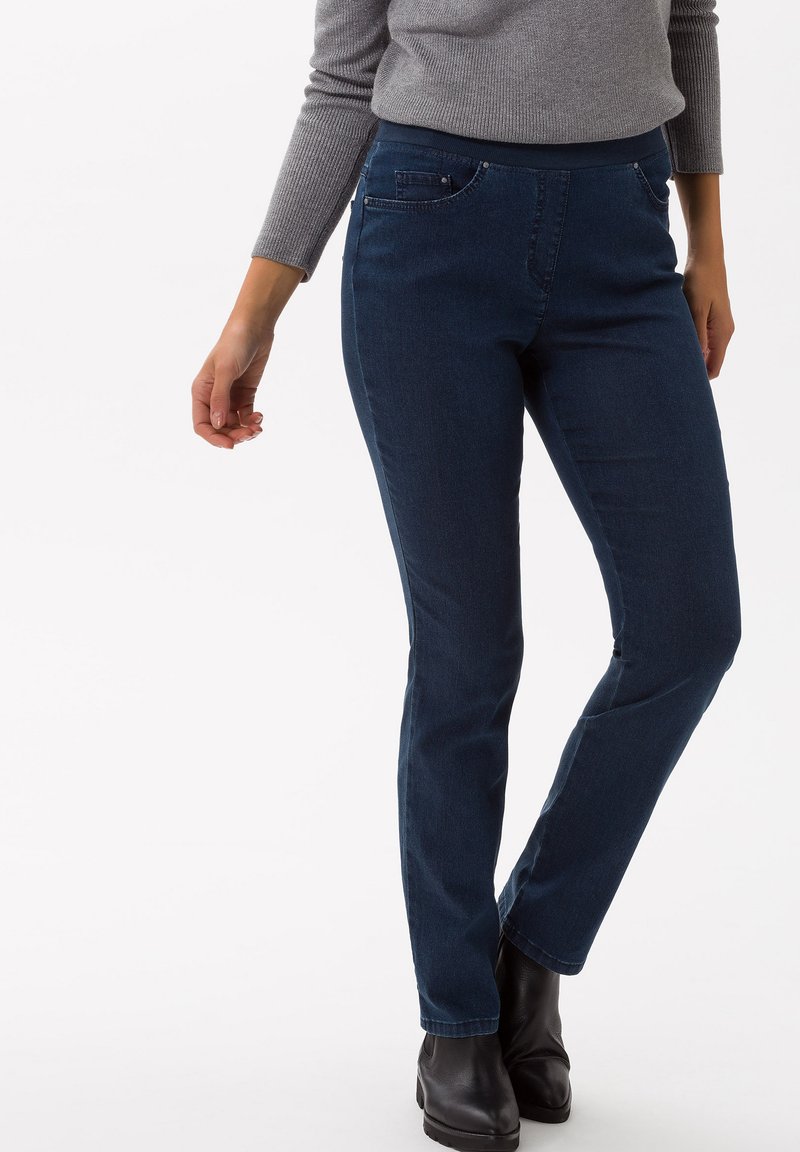 Jeans in denim blu scuro con vestibilità slim, dotati di tasche frontali, una texture liscia e una vita leggermente elasticizzata. Si indossano stivali neri alla caviglia.