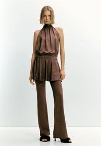 Ženska v brezrokavnem rjavem topu s halter krojem, oblečena s krilom v obliki zvona in usklajenimi oprijetimi hlačami, dopolnjena z visokimi črnimi krznjenimi petkami.