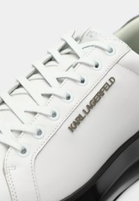 Tenisii din piele albă, cu o textură netedă, având șireturi de culoare albastru deschis și un logo metalic "KARL LAGERFELD" pe laterale. Talpă neagră.