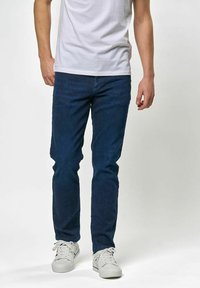 Tumši zili džinsa džinsi ar slaidu dizainu, pieciem kabatām un rāvējslēdzēja aizdari, kopā ar baltām sporta kurpēm un vienkāršu baltu T-kreklu.