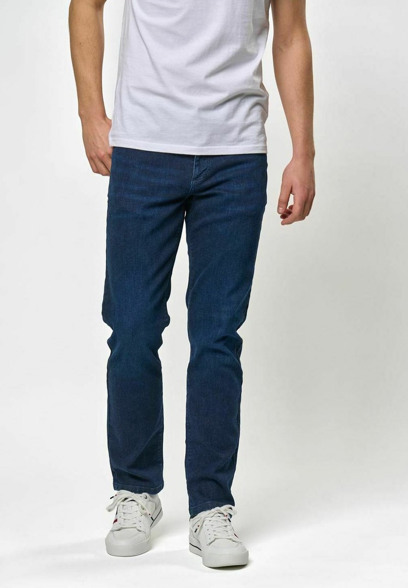 Tumši zili džinsa džinsi ar slaidu dizainu, pieciem kabatām un rāvējslēdzēja aizdari, kopā ar baltām sporta kurpēm un vienkāršu baltu T-kreklu.