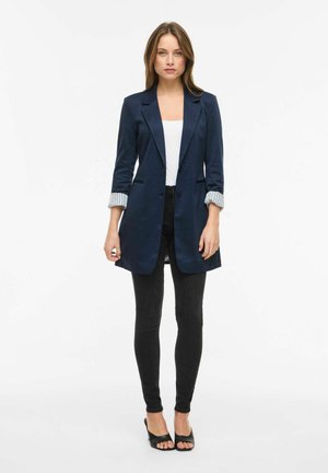 Kobieta stojąca w granatowej marynarce z podwiniętymi rękawami, białym topie, czarnych obcisłych spodniach i czarnych sandałkach na obcasie na tle jednolitej białej ściany.