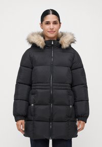 Mujer vestida con un abrigo de invierno negro acolchado con capucha en piel sintética, cierre frontal con cremallera y bolsillos con botones a presión, de pie contra un fondo liso.
