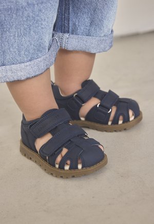 Kinder-Sandalen in Marineblau aus weichem Material, mit mehreren Riemen, offenen Zehen und einer rutschfesten braunen Sohle für Stabilität.