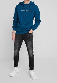 Blue hoodie with vertical stripes, embroidered text in white, paired with black distressed jeans featuring paint splatter, and white sneakers.