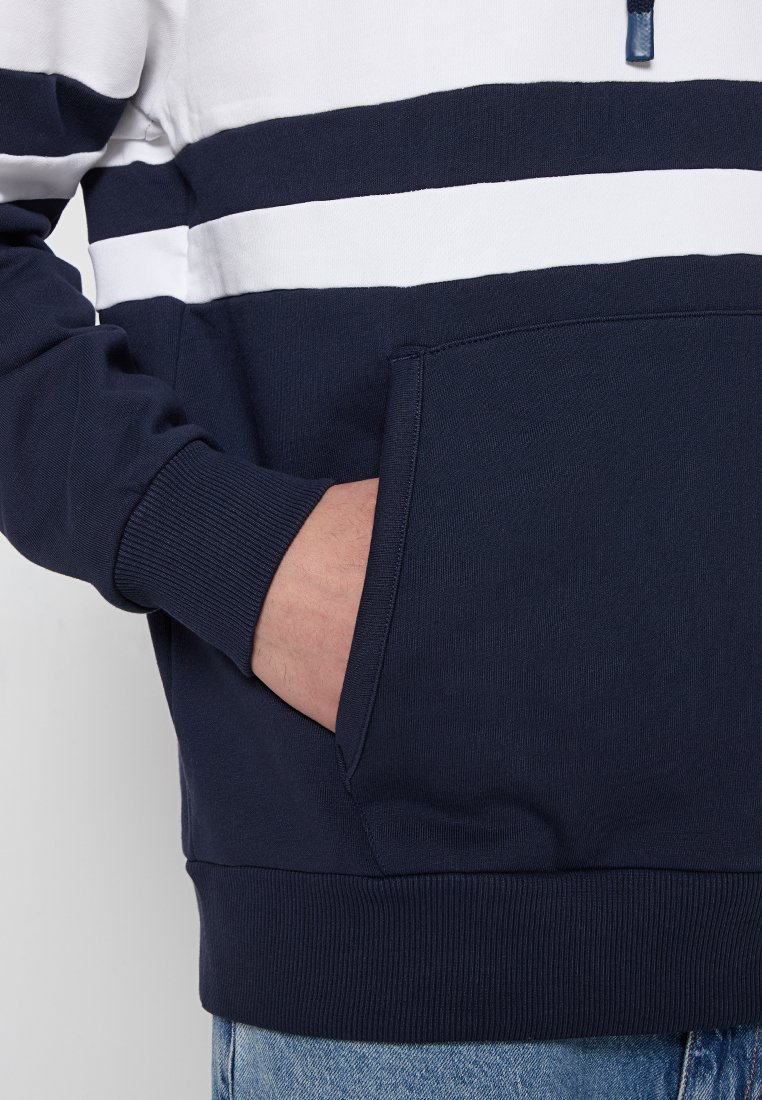 Primo piano di una persona che indossa un hoodie a righe blu e bianche con la mano nella tasca anteriore e jeans blu visibili.