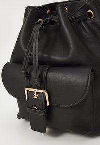 Sac seau en simili cuir noir avec une poche avant à rabat et boucle dorée, fermeture à cordon, texture lisse avec un minimalisme dans les détails.