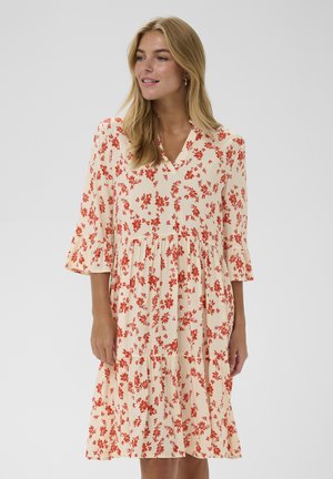 Žena s blond vlasmi, oblečená v krémových šatách po kolená s oranžovým kvetinovým vzorom a trojštvrťovými rukávmi s volánmi.