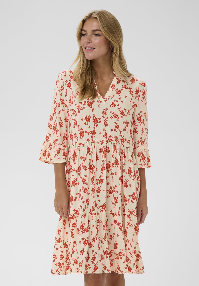 Kvinde med blondt hår iført en cremefarvet knælængde kjole med orange blomsterprint og trekvartlange ærmer med flæser.