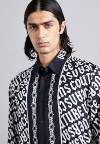 Νέος άνδρας με μακριά σκούρα μαλλιά που φοράει ένα μαύρο και λευκό πουκάμισο με έντονο γοτθικό κείμενο και σχέδια αλυσίδας στο εμπρός μέρος.