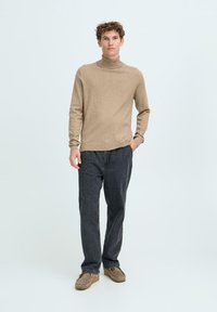 Beige garbópulóver puha anyagból, sötétszürke bő szárú nadrággal és bézs papuccsal párosítva, világos háttér előtt.
