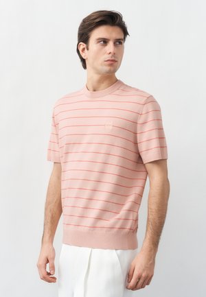 Νέος άνδρας που φοράει ανοιχτό ροζ πουλόβερ με κοντά μανίκια και λεπτές κόκκινες οριζόντιες ρίγες, καθώς και λευκό παντελόνι, κοιτάζει προς τα αριστερά του.