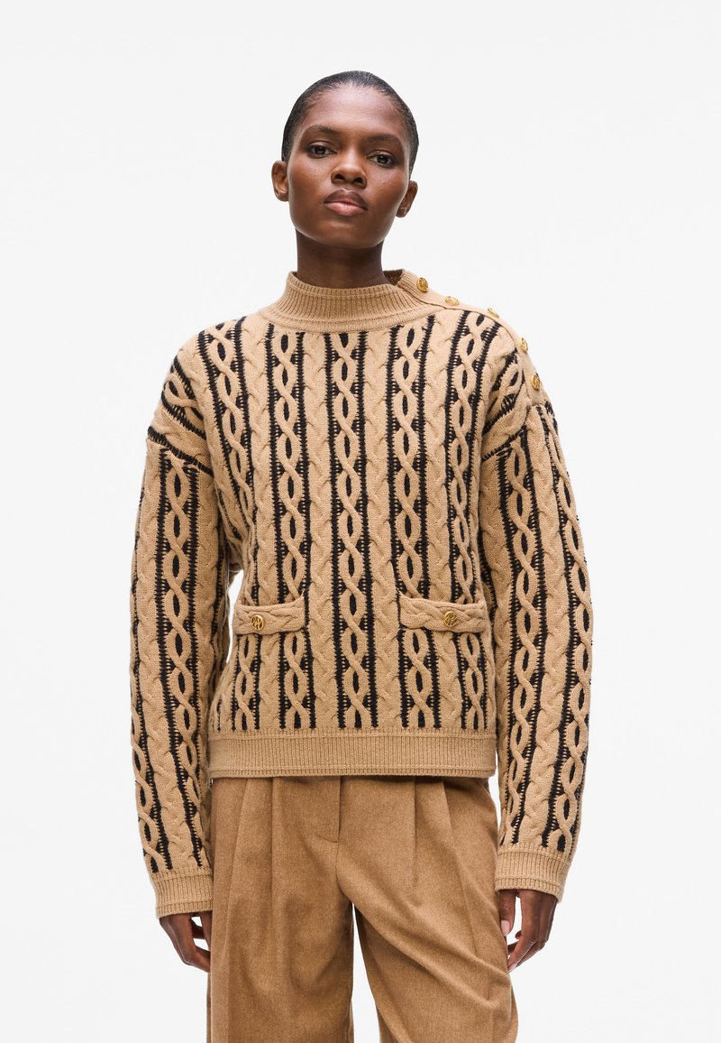 Pulover bej din tricot cu modele negre în formă de cablu, guler înalt, două buzunare frontale și accente cu nasturi aurii pe umeri.