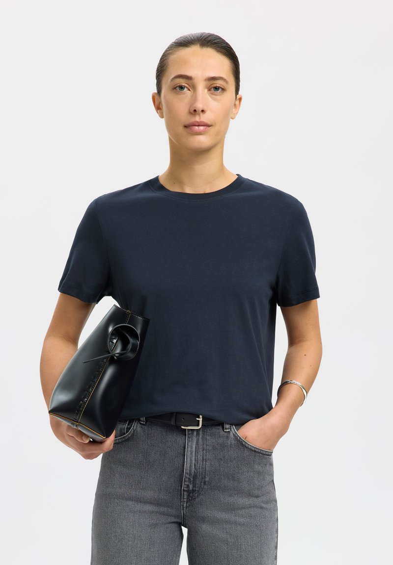 Mujer con el pelo recogido, vistiendo una camiseta oscura y vaqueros grises, sosteniendo un bolso de mano negro de cuero sobre un fondo liso.
