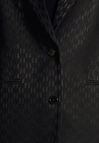 Blazer noir texturé avec un motif géométrique subtil, deux boutons fermés et deux poches passepoilées visibles à l'avant.