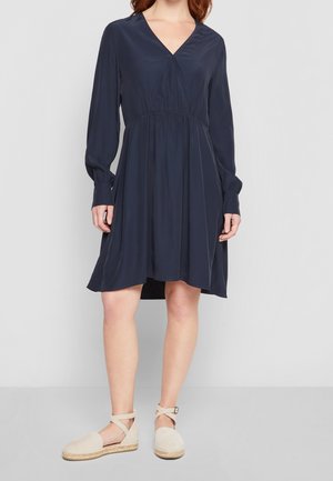 Vrouw draagt een marineblauwe jurk met lange mouwen en V-hals en beige espadrilles met enkelbandjes, staand tegen een effen achtergrond.