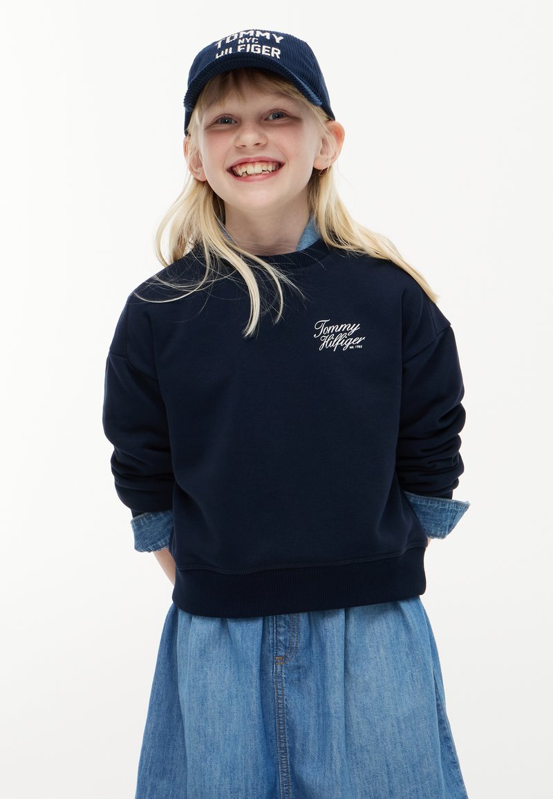 Fille souriante aux longs cheveux blonds porte une casquette et un sweat-shirt Tommy Hilfiger bleu marine sur une robe en jean bleue, les mains derrière le dos sur fond blanc.