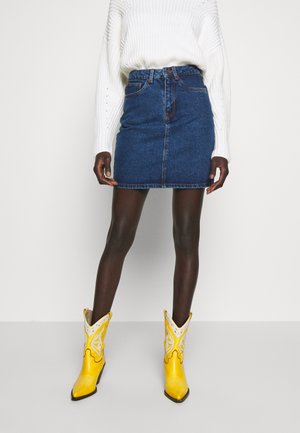 Dunkelhäutige Person trägt einen weißen Strickpullover, einen blauen Jeansminirock und leuchtend gelbe Cowboy-Stiefel und steht vor einem einfarbigen Hintergrund.