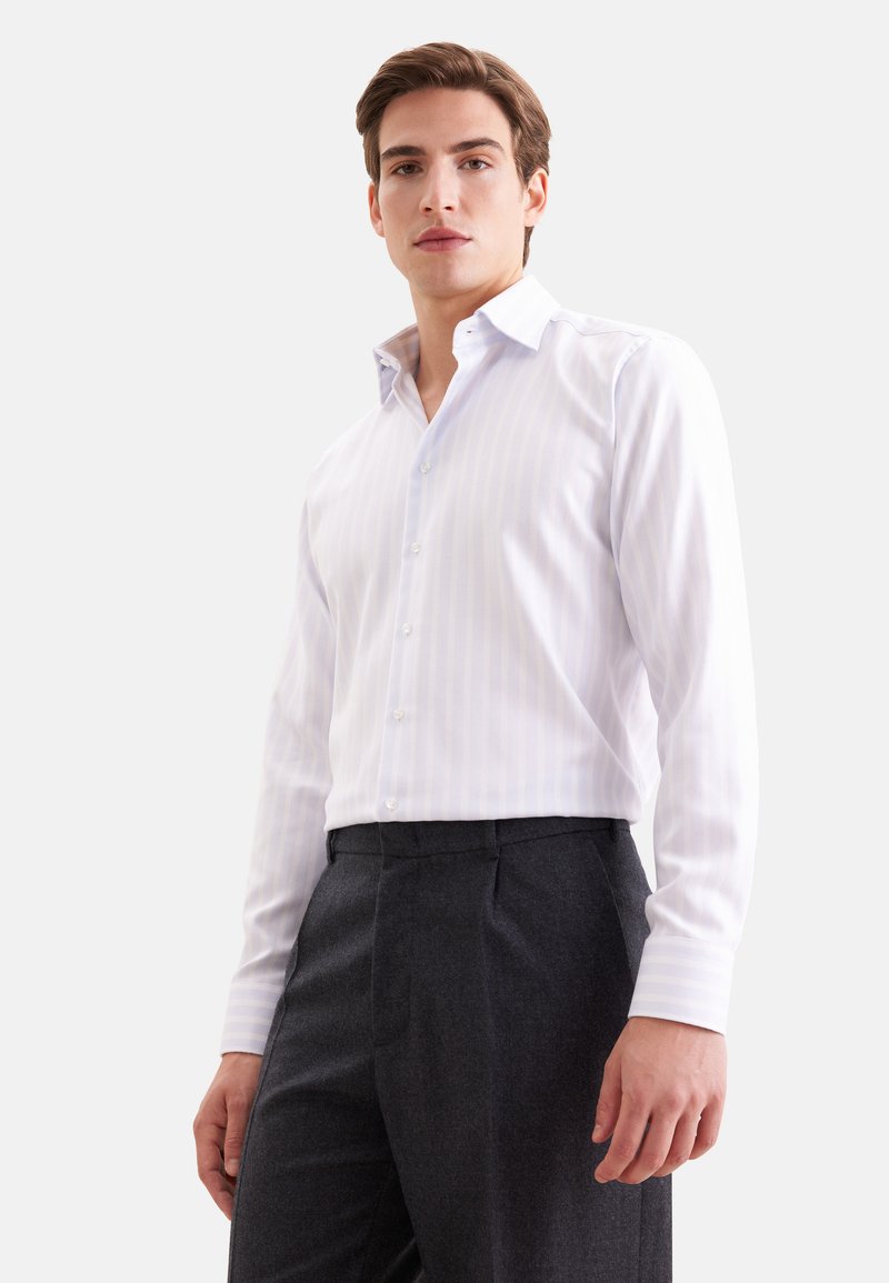 Jeune homme portant une chemise blanche à manches longues boutonnée et un pantalon gris foncé, debout devant un fond clair uni.