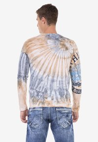 Langärmliges Batikshirt in Beige, Blau und Grau; mit Rundhalsausschnitt und grafischen Motiven auf den Ärmeln. Kombiniert mit ausgewaschenen blauen Jeans.
