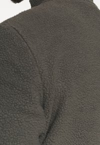 Mężczyzna w ciemnoszarej, strukturalnej kurtce polarowej, pokazujący górną część pleców i ramienia z widocznymi detalami szwu.