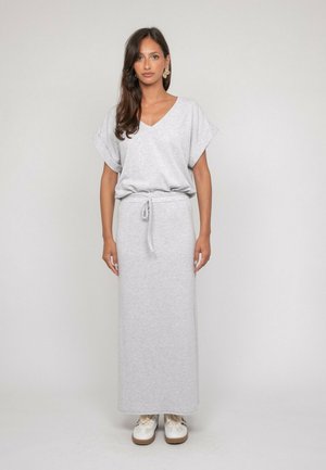Femme debout portant un haut gris clair à manches courtes et col en V avec une longue jupe assortie nouée à la taille et des baskets blanches.
